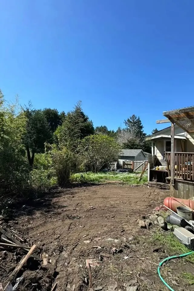 Backyard cleared to bare soil beside a deck and tree line