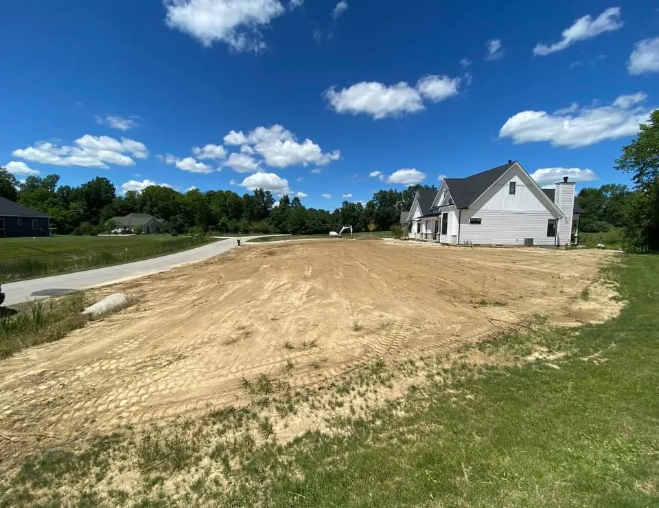 Lot graded to bare earth beside a new house with an excavator in the distance
